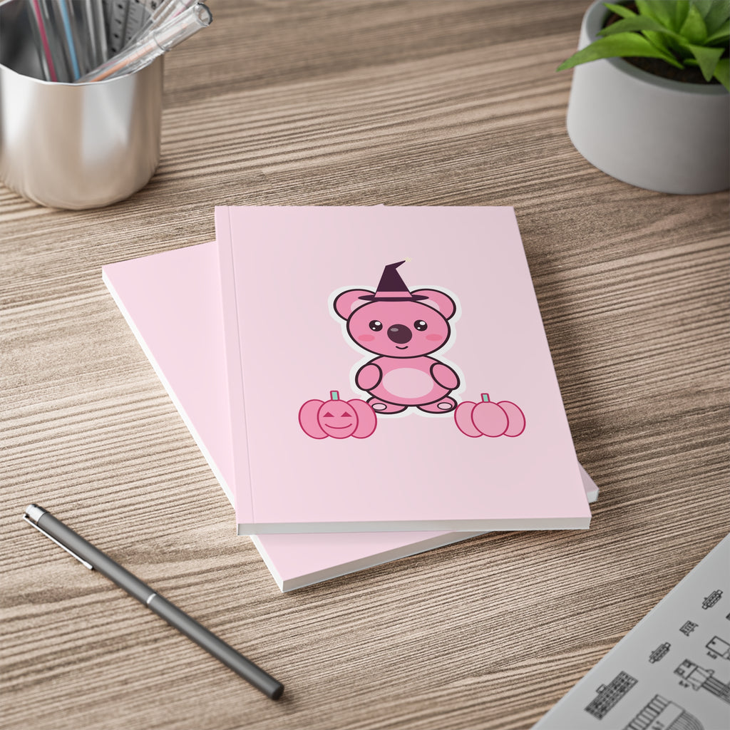 Pink A5 notebook with witch-hat Koa and pink pumpkins on a wooden desk